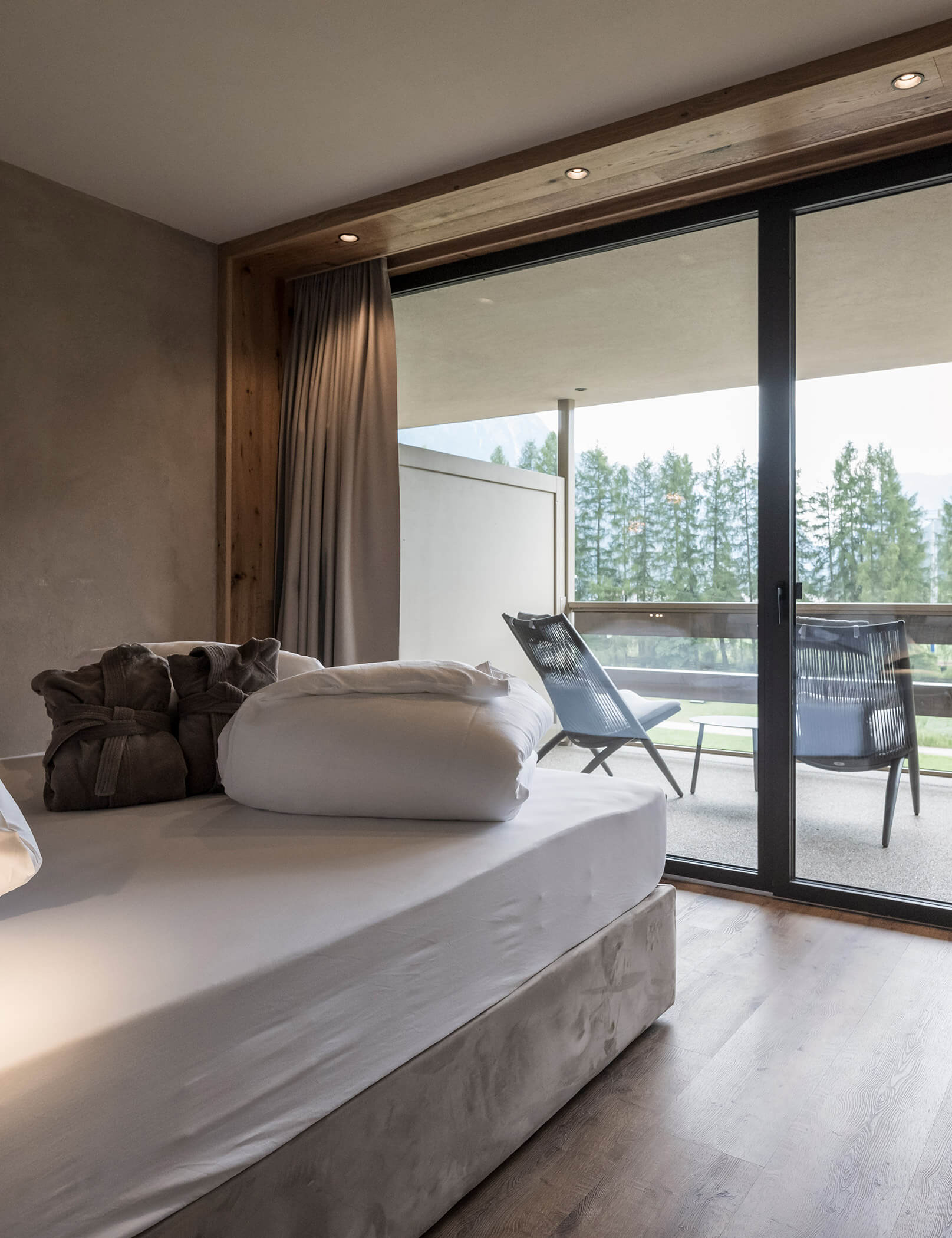 Letto matrimoniale con piumone bianco e accappatoi marroni, balcone con sedie sullo sfondo - Hotel Patris