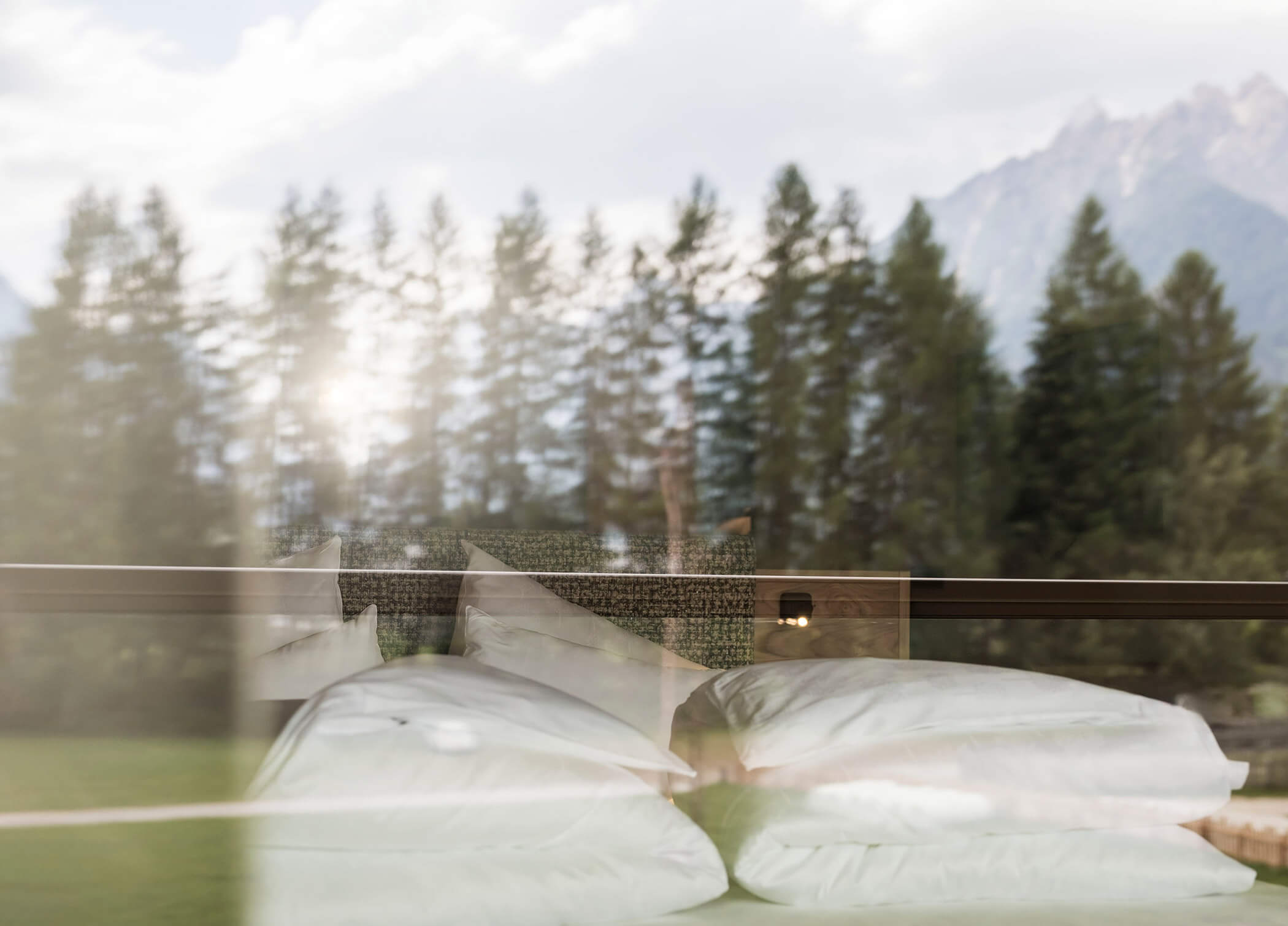 Letto matrimoniale con coperte bianche, alberi e montagne di Dobbiaco riflessi nel vetro della finestra - Hotel Patris